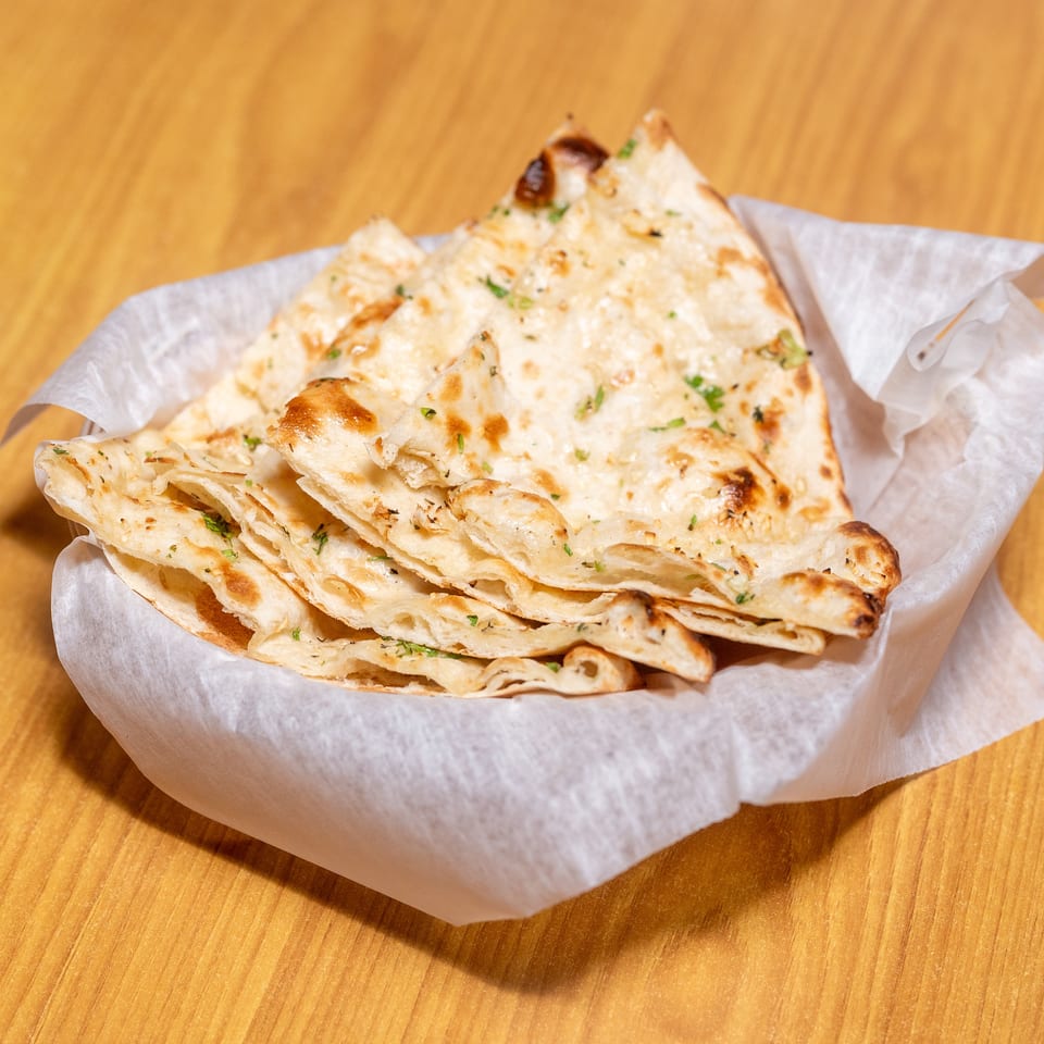 Stack of golden, flaky naan breads sprinkled with herbs, served in a paper-lined basket on a wooden table.
