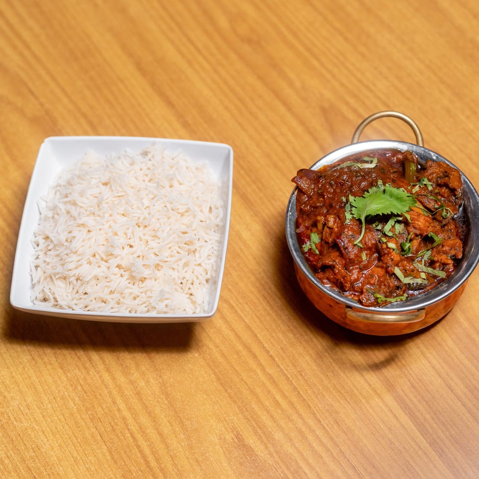 Basmati rice served beside a garnished red curry in a copper-handled bowl on a wooden table.