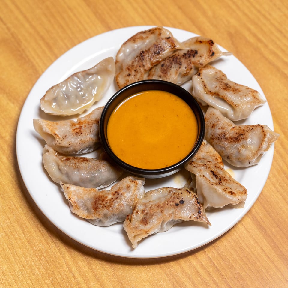 Pan-fried dumplings arranged around a bowl of orange dipping sauce on a white plate atop a wooden table.
