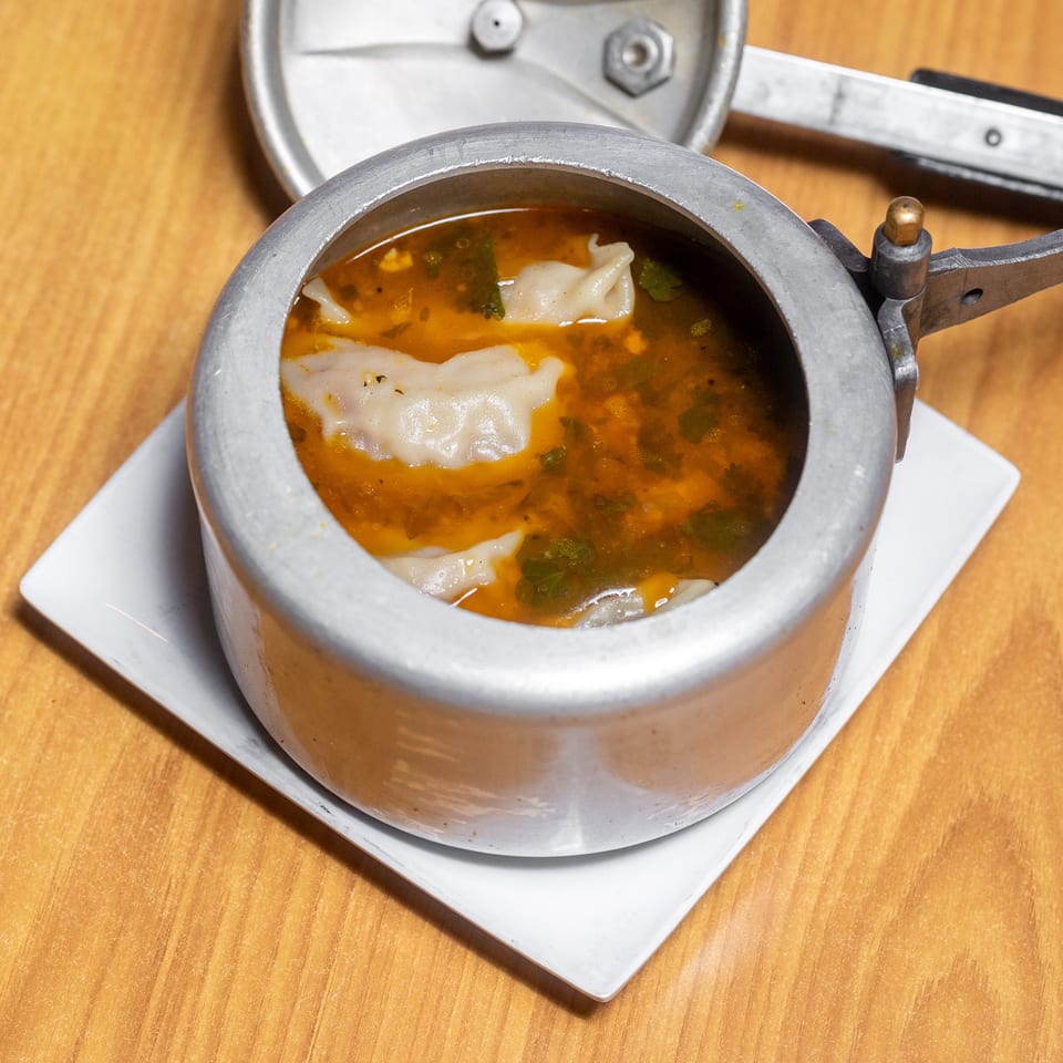 Open metal serving pot filled with dumplings in orange herb broth on a white plate atop a wooden table.