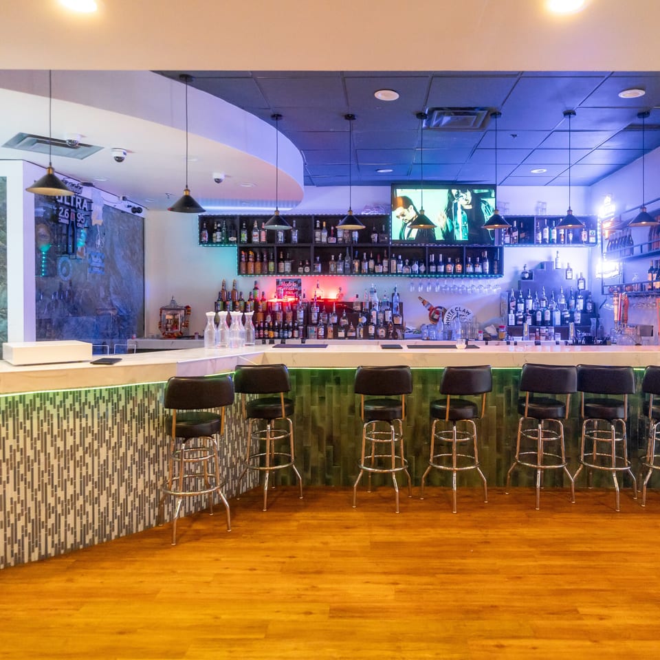 A brightly lit restaurant bar displays liquor bottles, pendant lights, colorful accents, and black stools along a tiled counter.