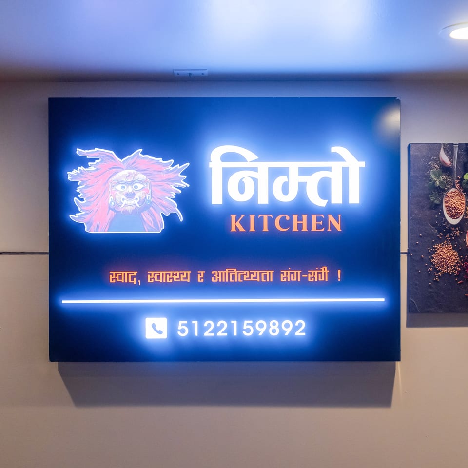 Illuminated blue sign for Swad Kitchen featuring a colorful traditional mask logo, Nepali text, and phone number.