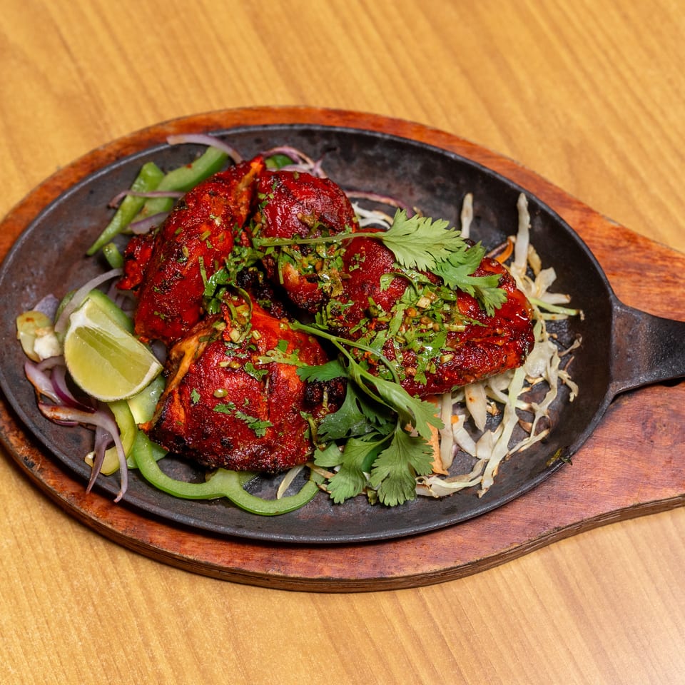Red grilled chicken pieces garnished with cilantro, served with peppers, onions, cabbage, and lime on a sizzling platter.