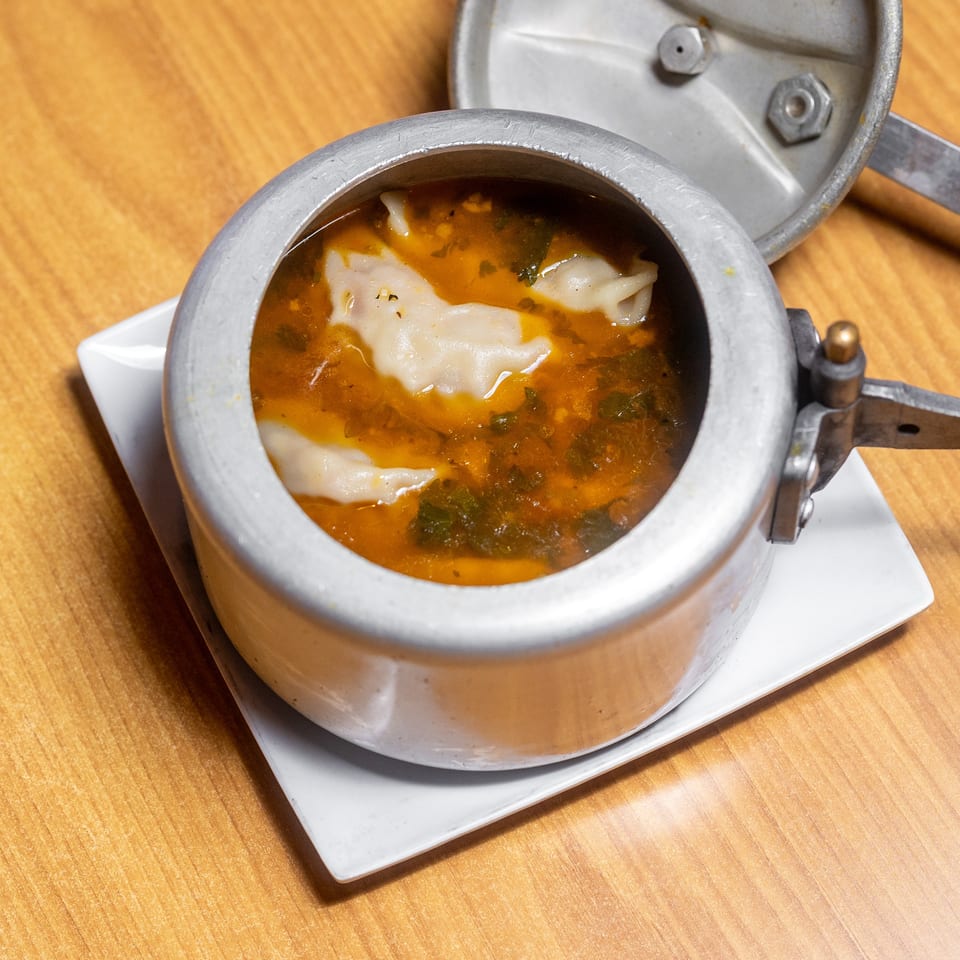Open metal serving pot filled with dumplings in orange herb broth on a white plate atop a wooden table.