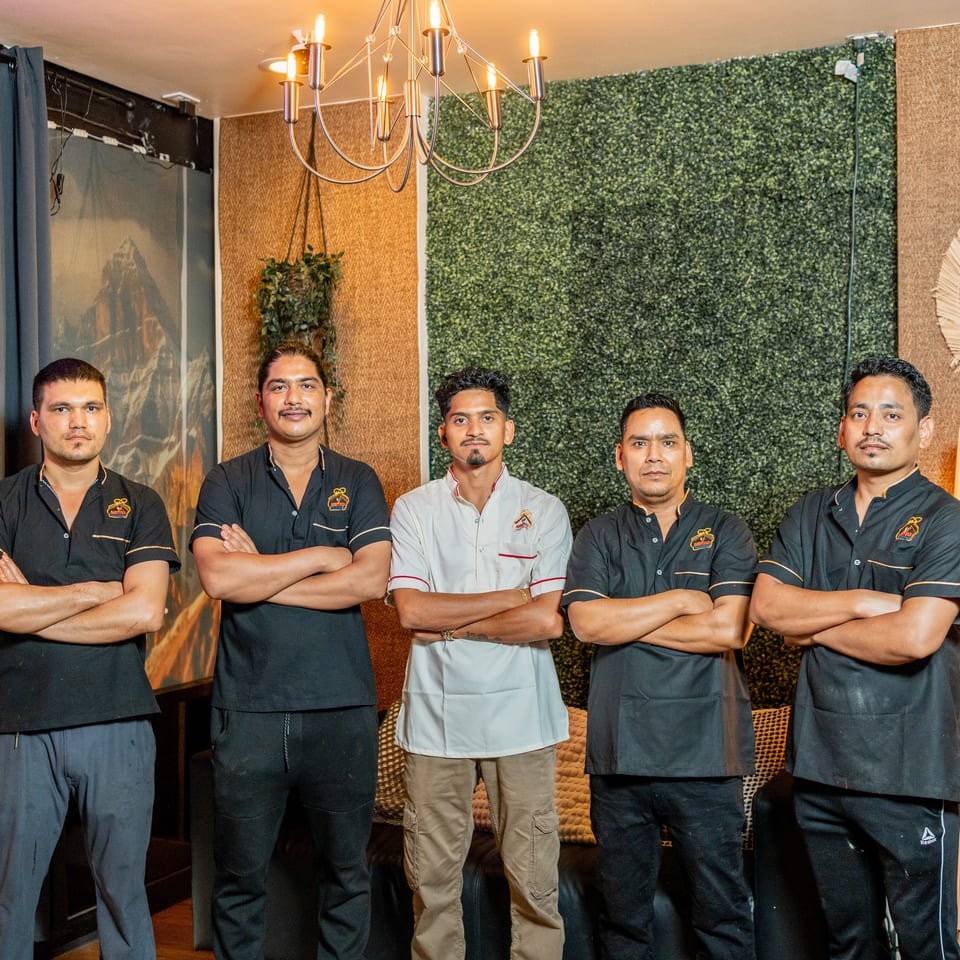 Five restaurant staff members pose with folded arms beneath a chandelier in a warmly decorated dining room.