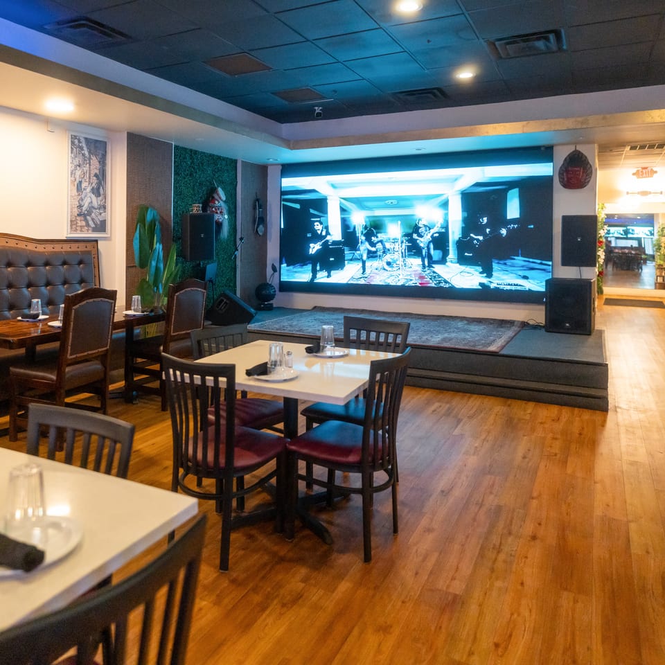 Restaurant dining room with set tables, leather booth seating, wood floors, and a large screen displaying a live band performance.