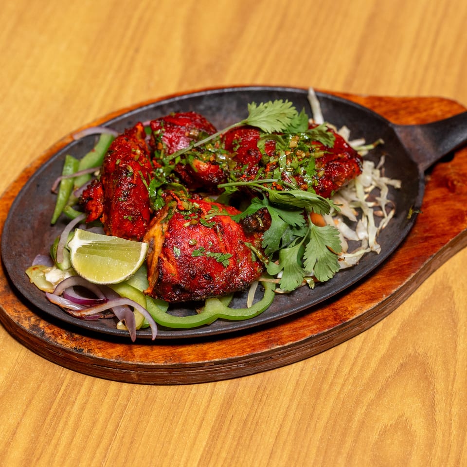 Red grilled chicken pieces garnished with cilantro, lime, onions, and peppers on a sizzling platter.
