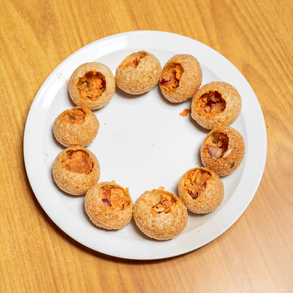 Crispy pani puri shells filled with spiced potato mixture arranged on a white plate atop a wooden table.