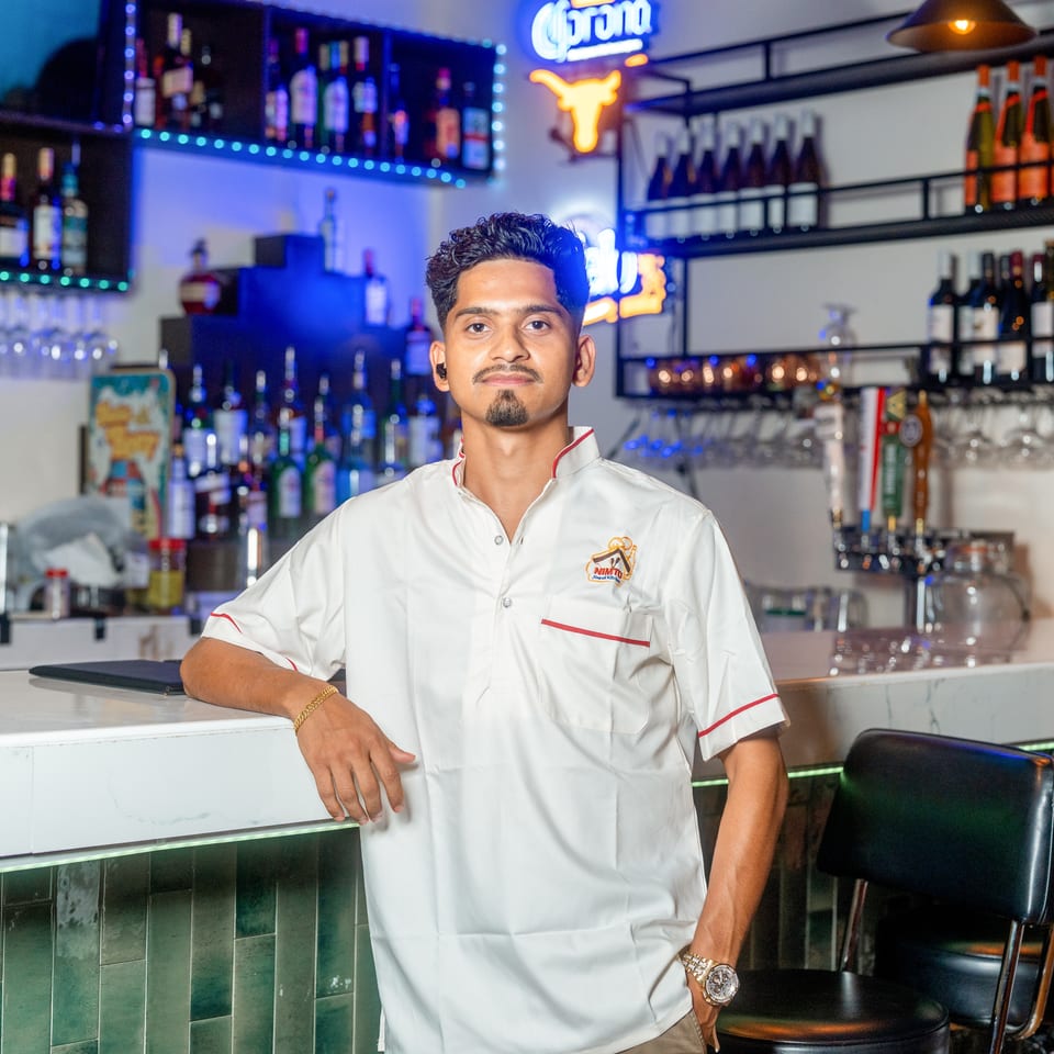 Restaurant staff member in a white uniform stands at a colorful, well-stocked bar beneath illuminated Corona signage.