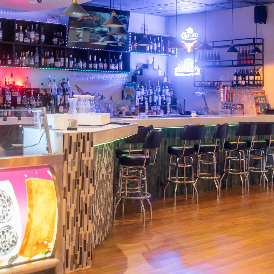 Colorfully lit restaurant bar stocked with liquor bottles, neon beer signs, pendant lights, and rows of black bar stools.