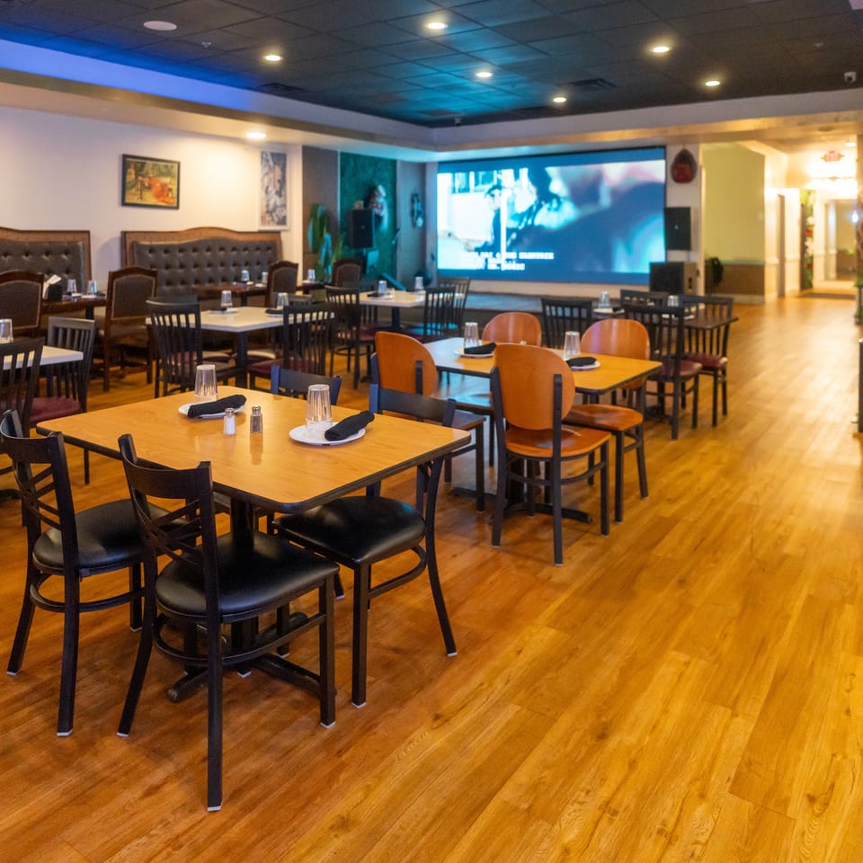 Spacious restaurant dining room with set tables, booth seating, polished wood floors, and a large projection screen.